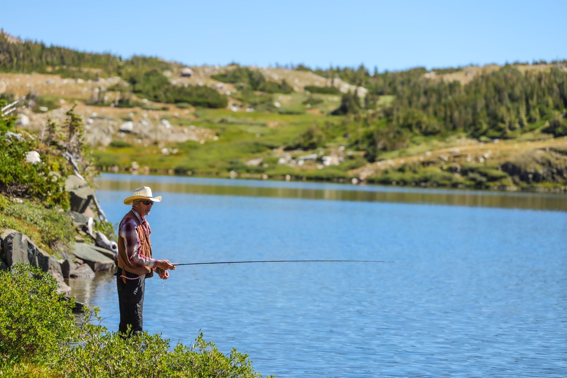 Person fishing from a rocky lakeshore beside a blue mountain lake under a clear sky