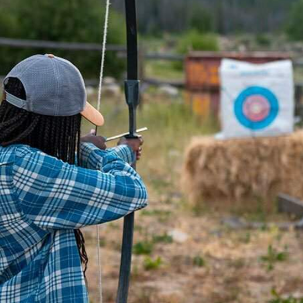 Person aiming a bow and arrow at an outdoor target near hay bales and grass.