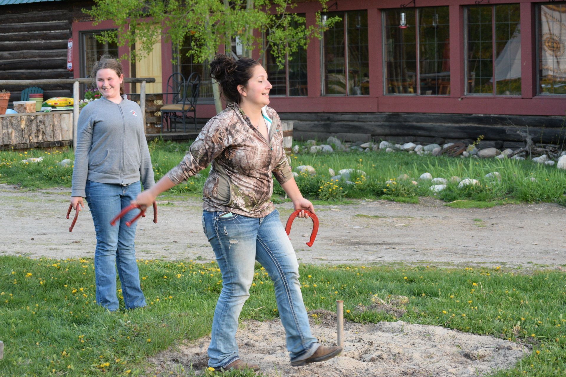 Two people outdoors tossing horseshoes near a rustic building