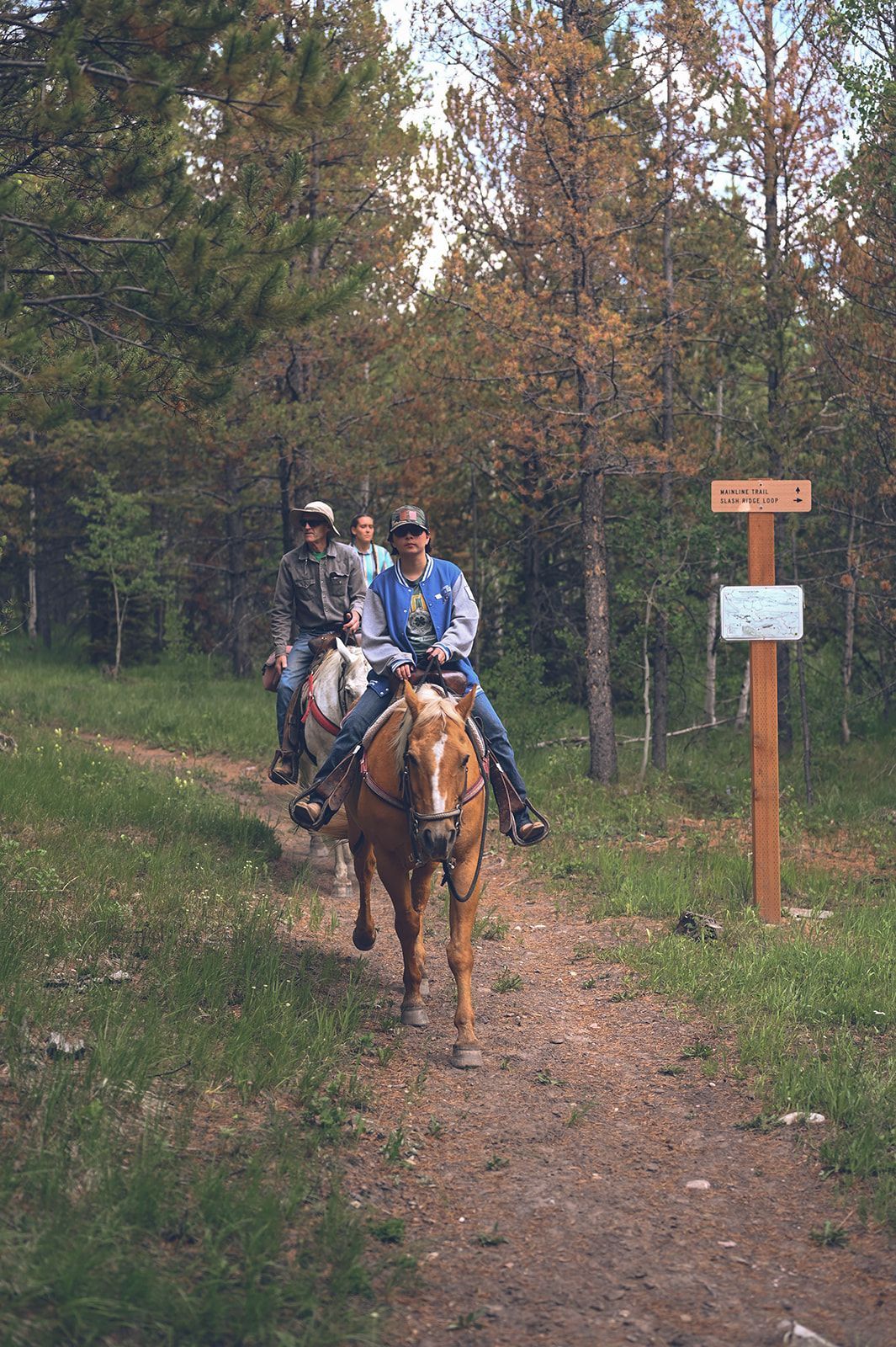 Two people horseback riding on a forest trail beside a wooden signpost.