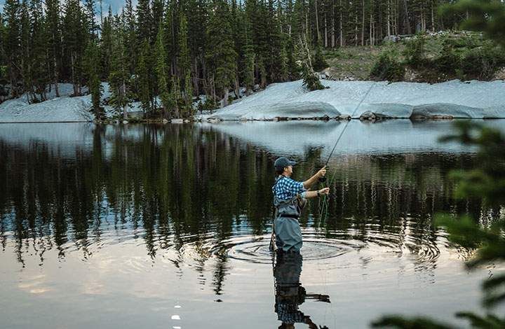 Person fishing in a calm forest lake, with snowy shoreline and tree reflections at dusk