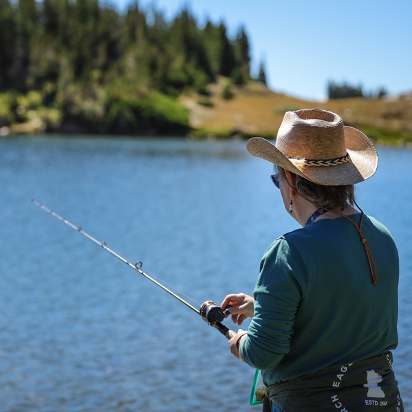 Person fishing from a lakeshore, wearing a straw hat, with forested hills in the background.
