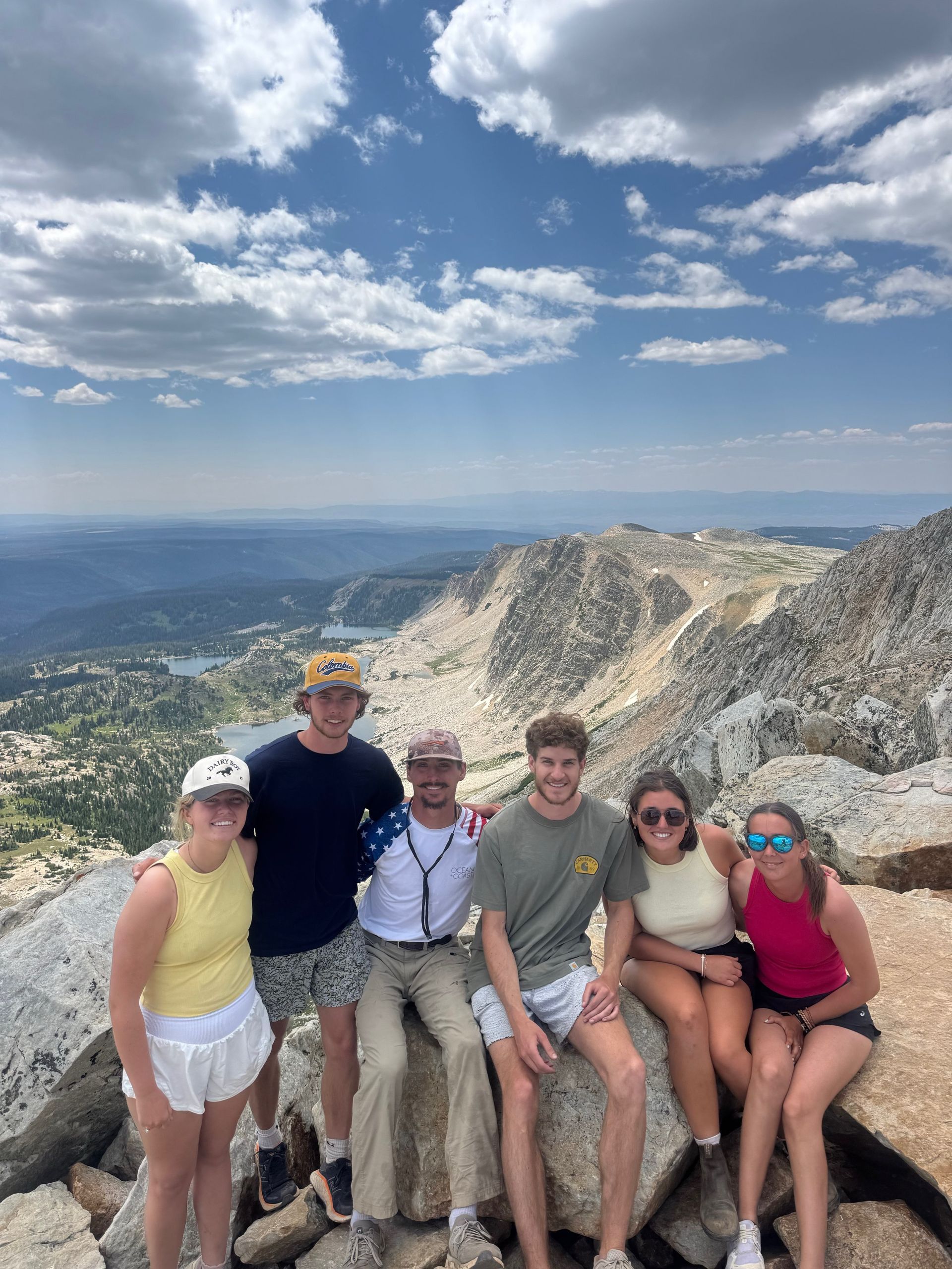 Five friends posing on a mountain overlook with rocky cliffs, blue sky, and a city in the distance