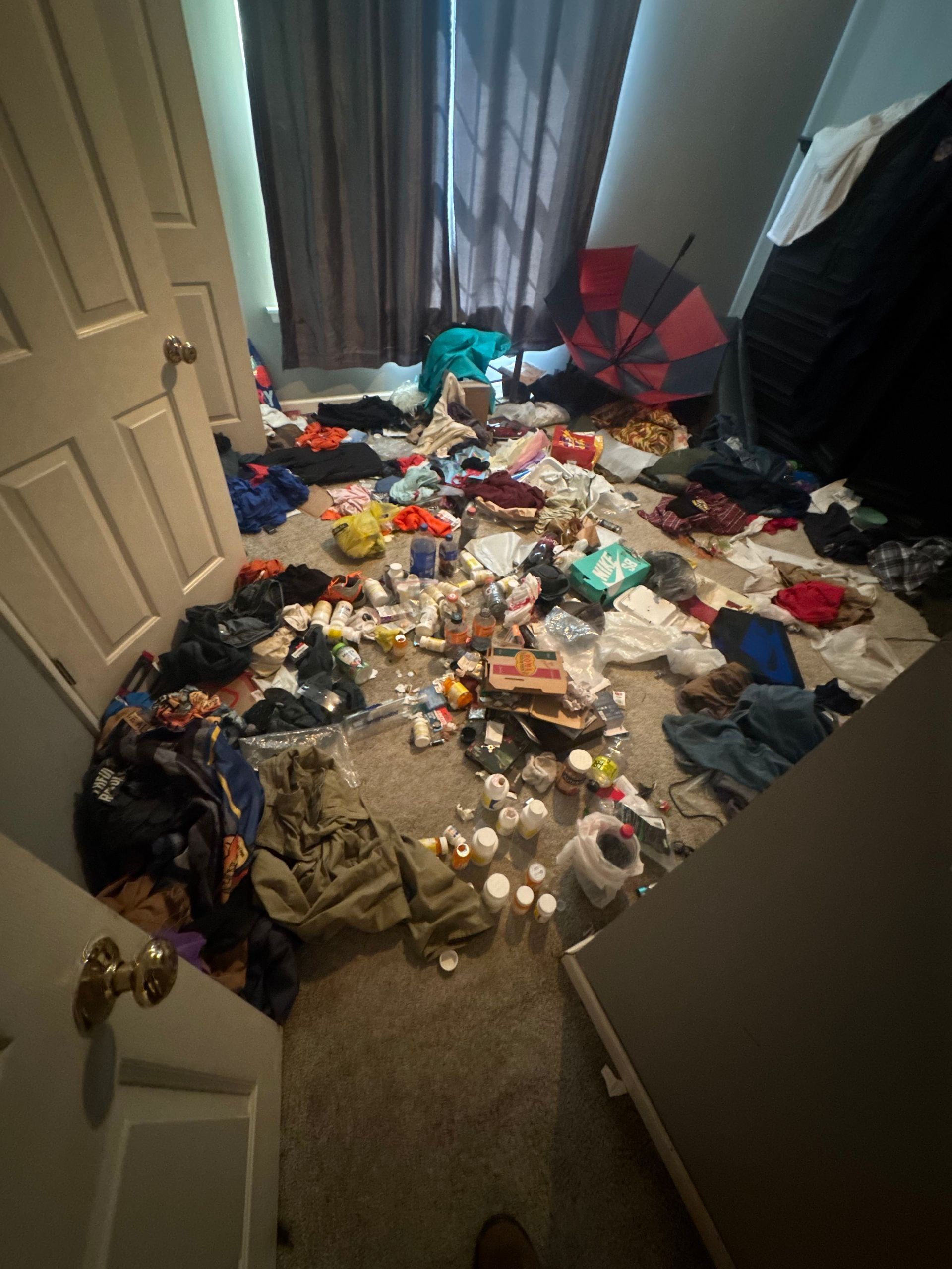 Cluttered bedroom floor covered with scattered clothes, toys, and trash near an open door and window curtain