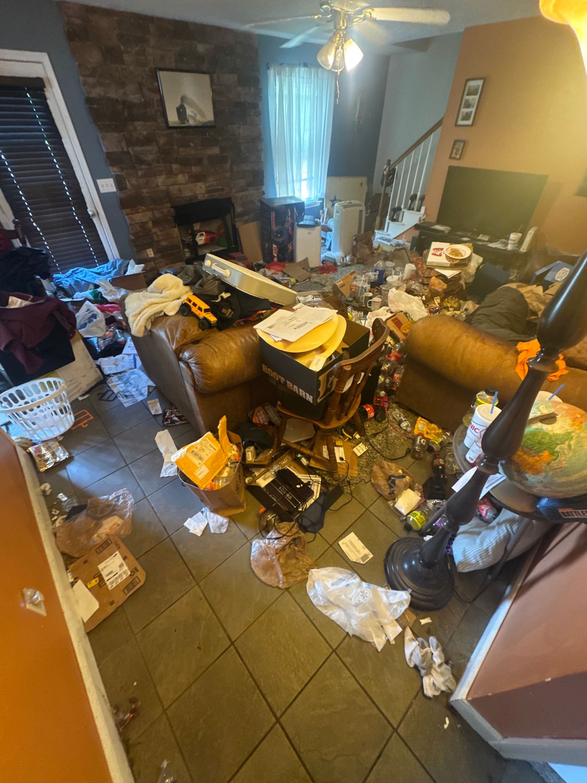 Cluttered living room with trash and bags scattered around a couch, tiled floor, and window light.