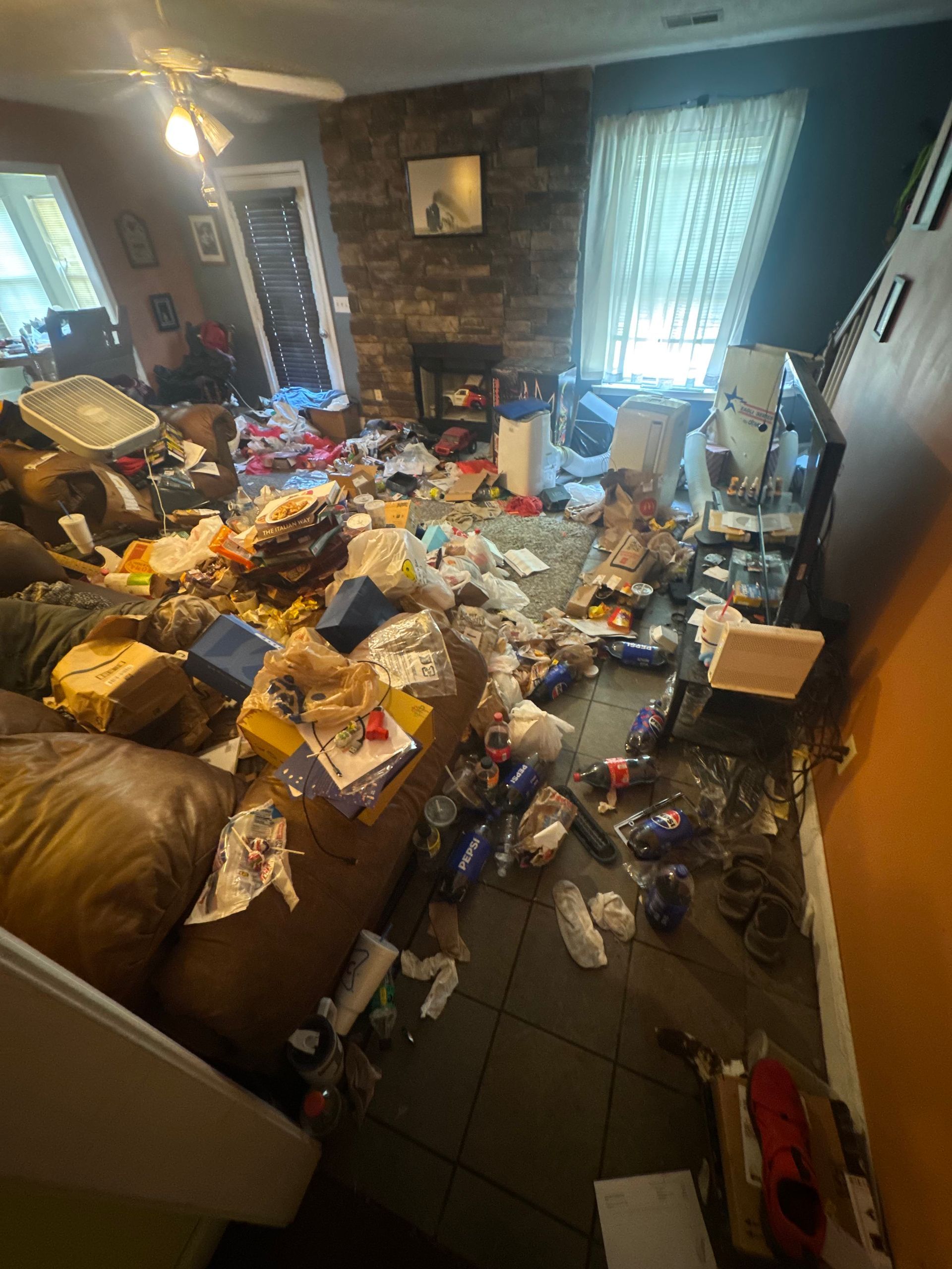 Cluttered living room with trash and debris covering the floor, couch, and tables, with sunlight from a window.