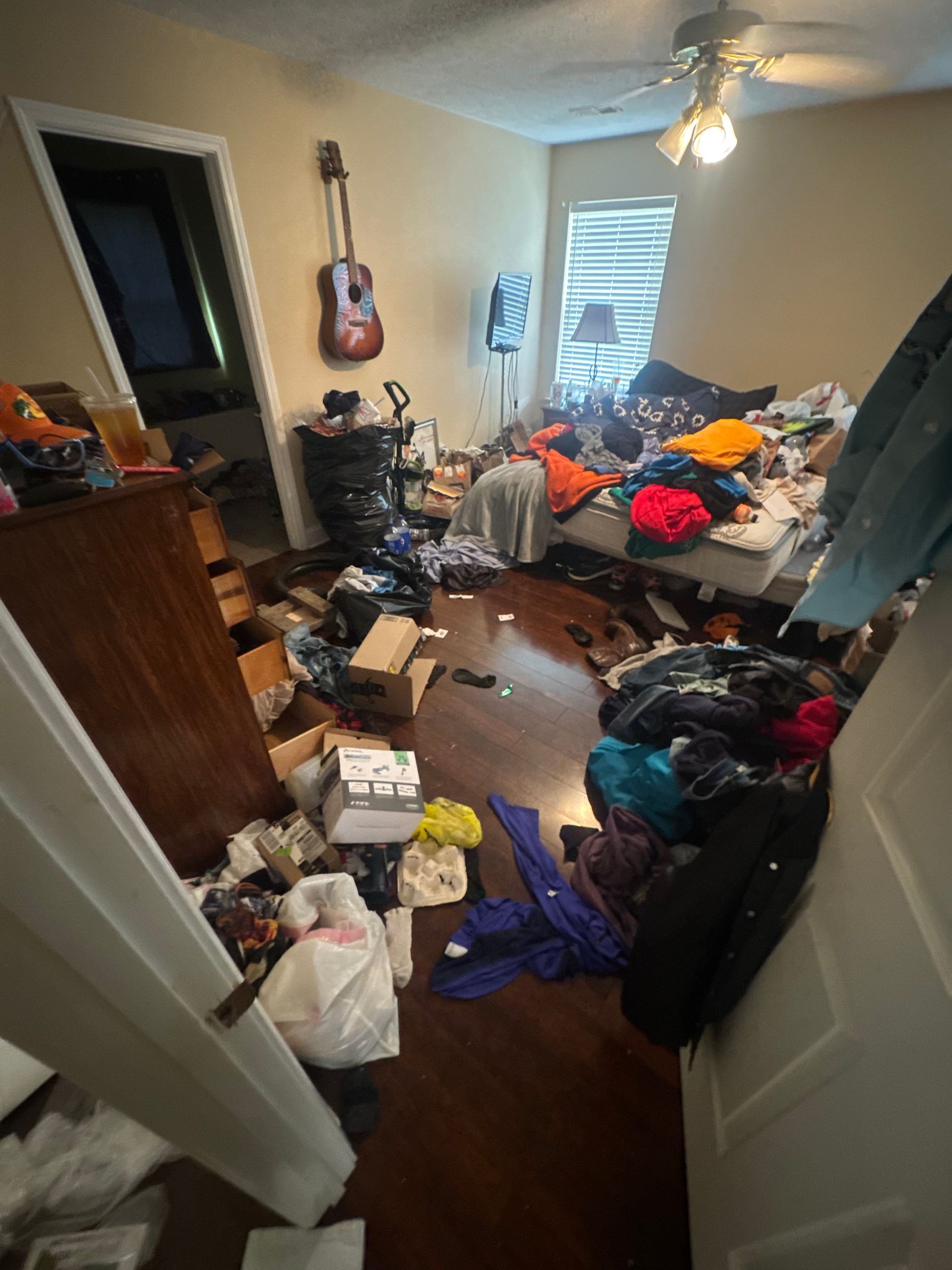 Cluttered bedroom with clothes and bags strewn across the floor and bed, daylight through a window.