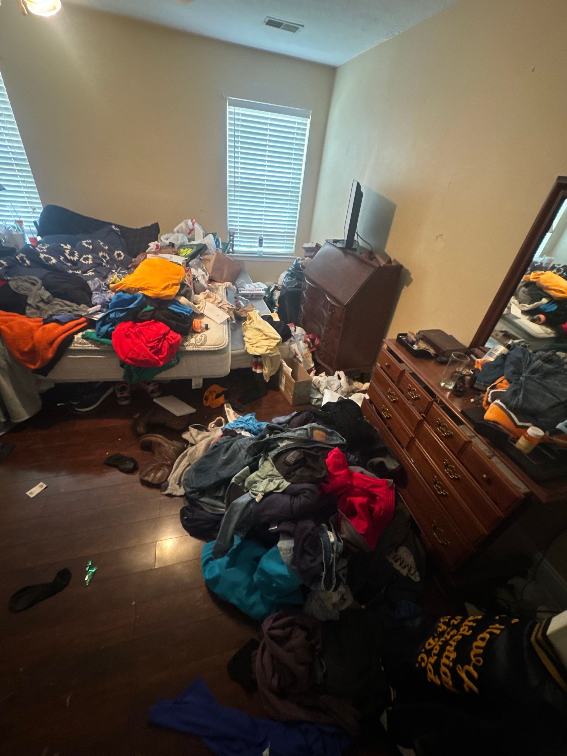 Cluttered bedroom with clothes piled on the bed, floor, dresser, and chair; TV against the wall.