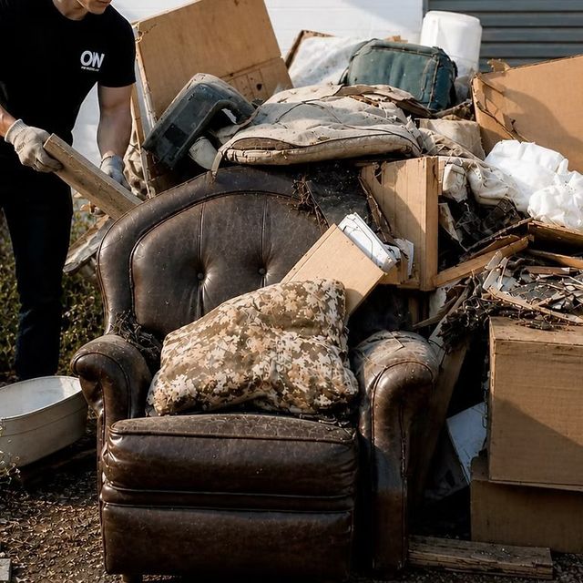 Armchair and household debris piled in a dumpster, with a person partly visible sorting through it.