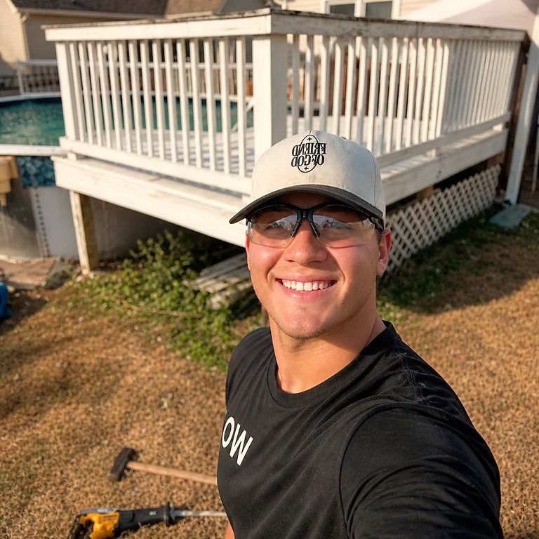 Smiling person taking a selfie in front of a white deck railing outdoors, holding a camera.