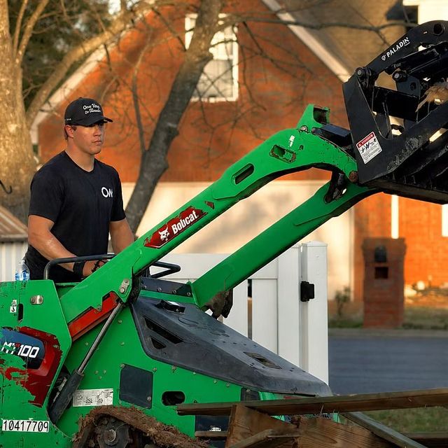 Man operating a green Bobcat skid steer outdoors near a brick building