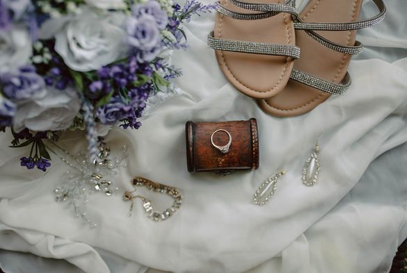Wedding accessories on white fabric: lavender bouquet, jeweled sandals, clutch, and sparkling jewelry.