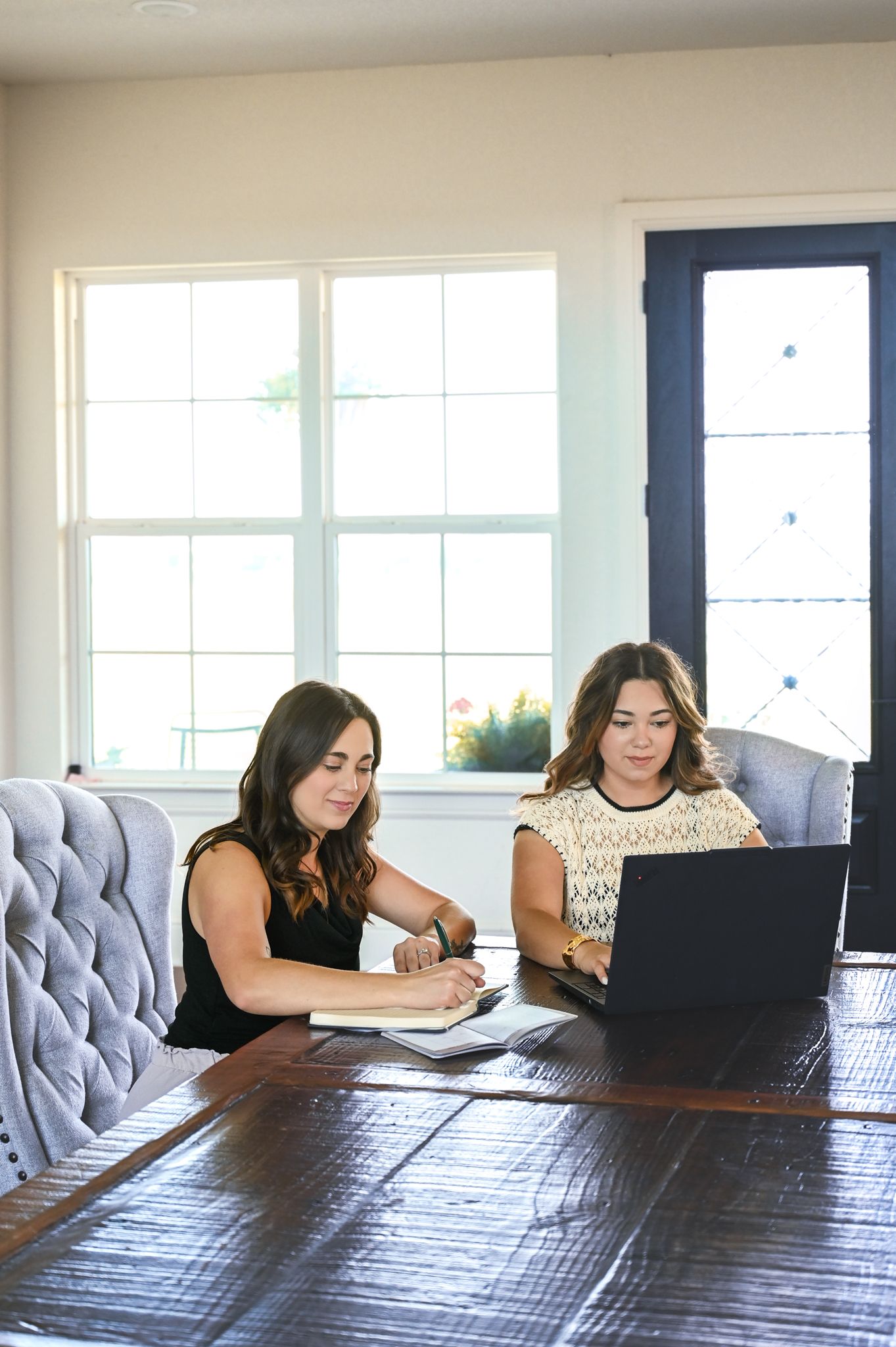 Two women sitting at a table, working on a laptop in a bright room