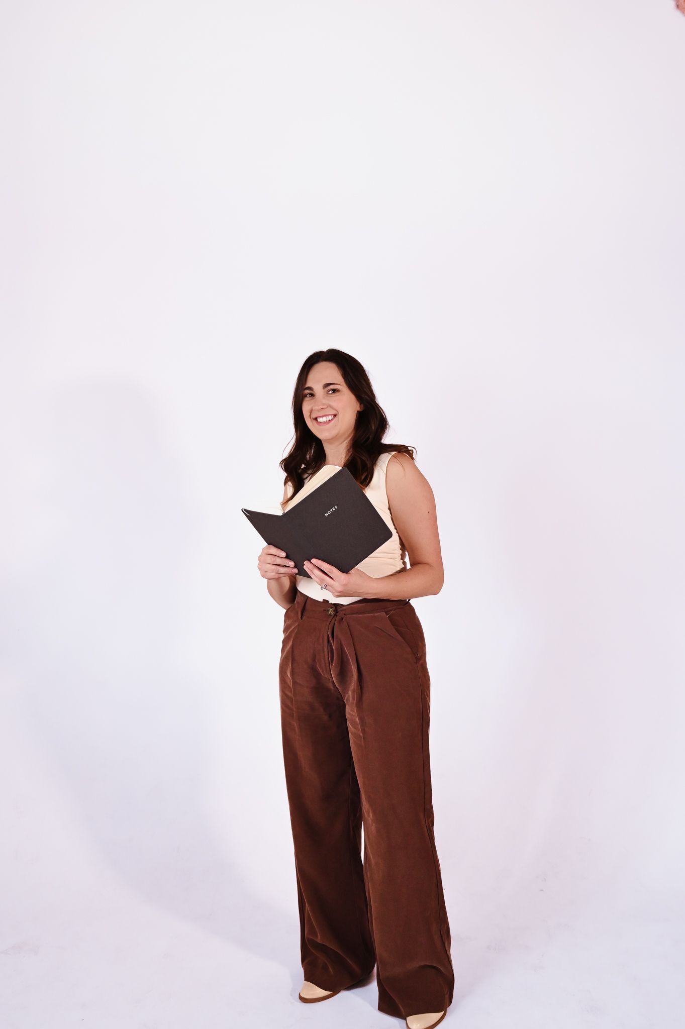 Woman in a pink top and brown pants holding a folder against a plain white background