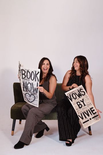 Two women seated on chairs holding bold typographic posters against a plain white background.