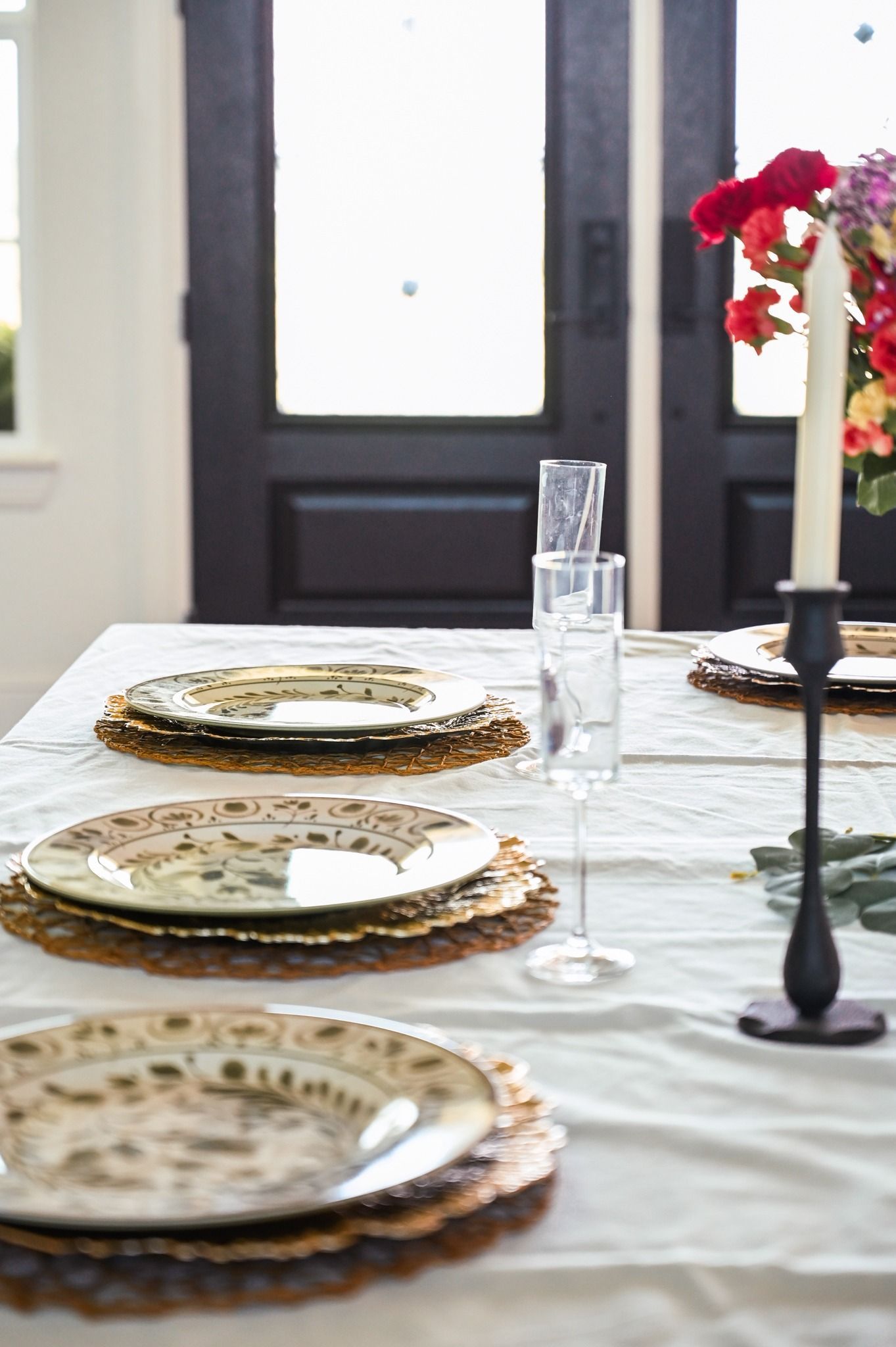 Set dining table with patterned plates, white tablecloth, and red flowers by a window