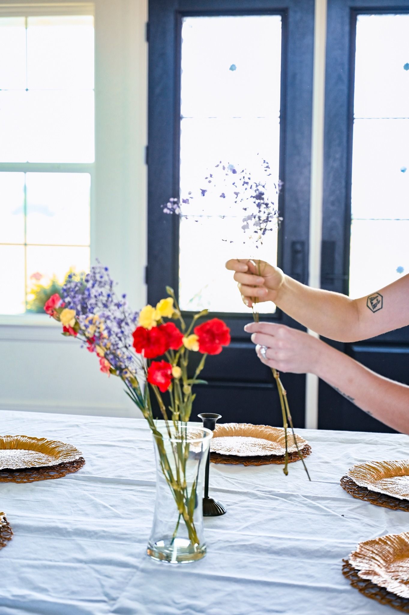 Hands setting a dining table with flowers and plates by a bright window
