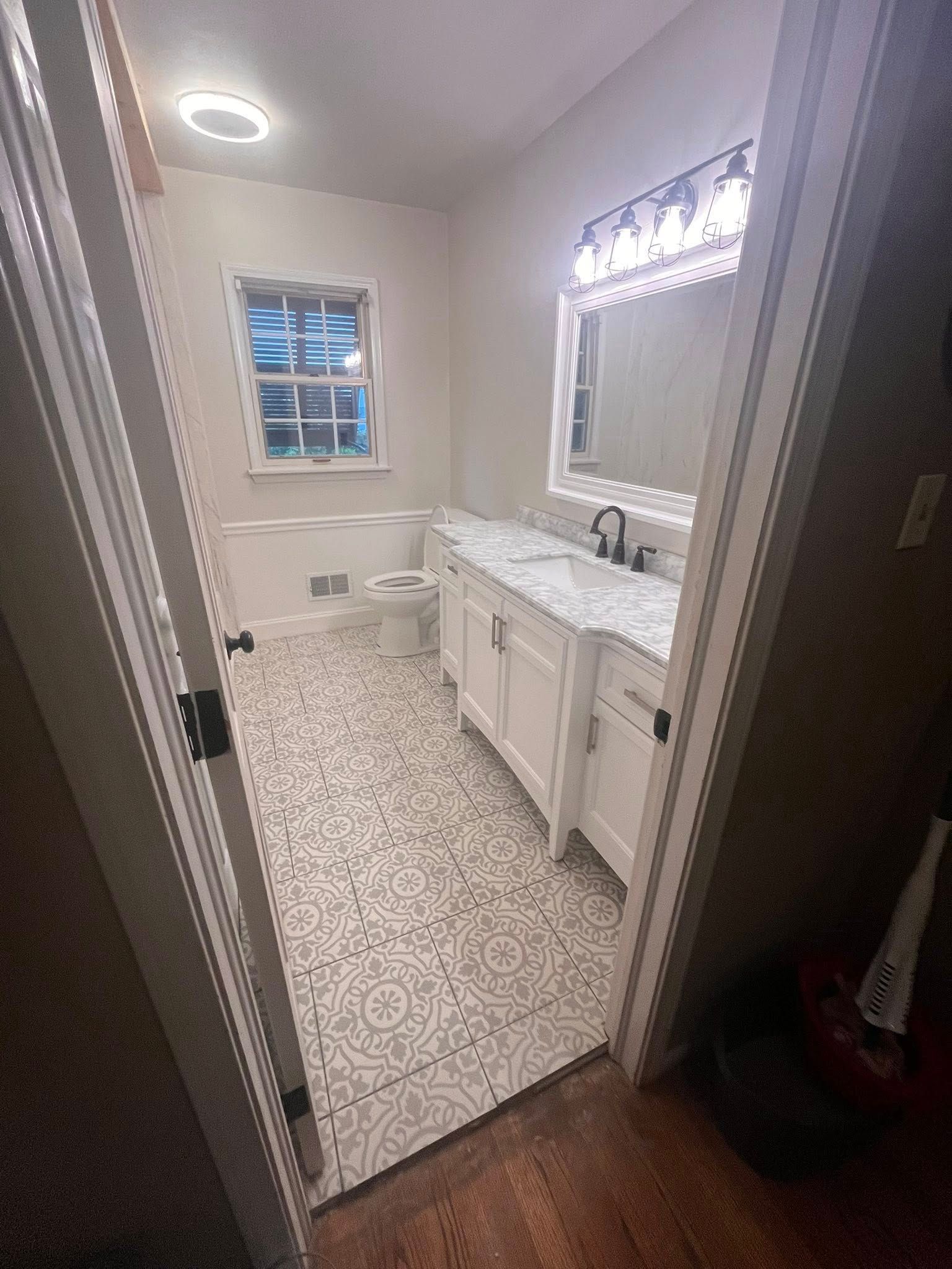 Small bathroom with patterned floor, white vanity, lit mirror, and window.