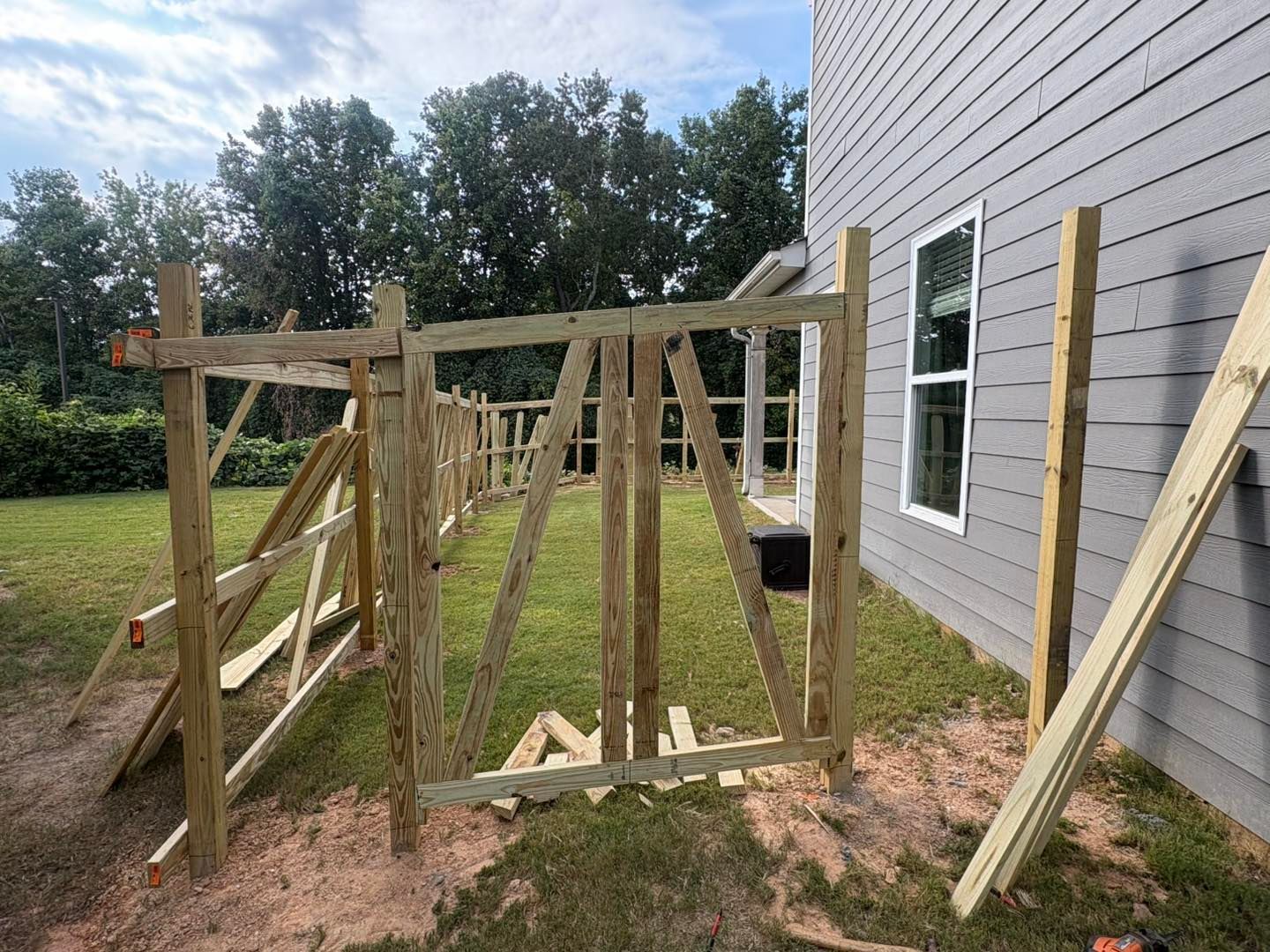 Wooden scaffold frame beside a house, with lumber leaning against the siding in a grassy yard.