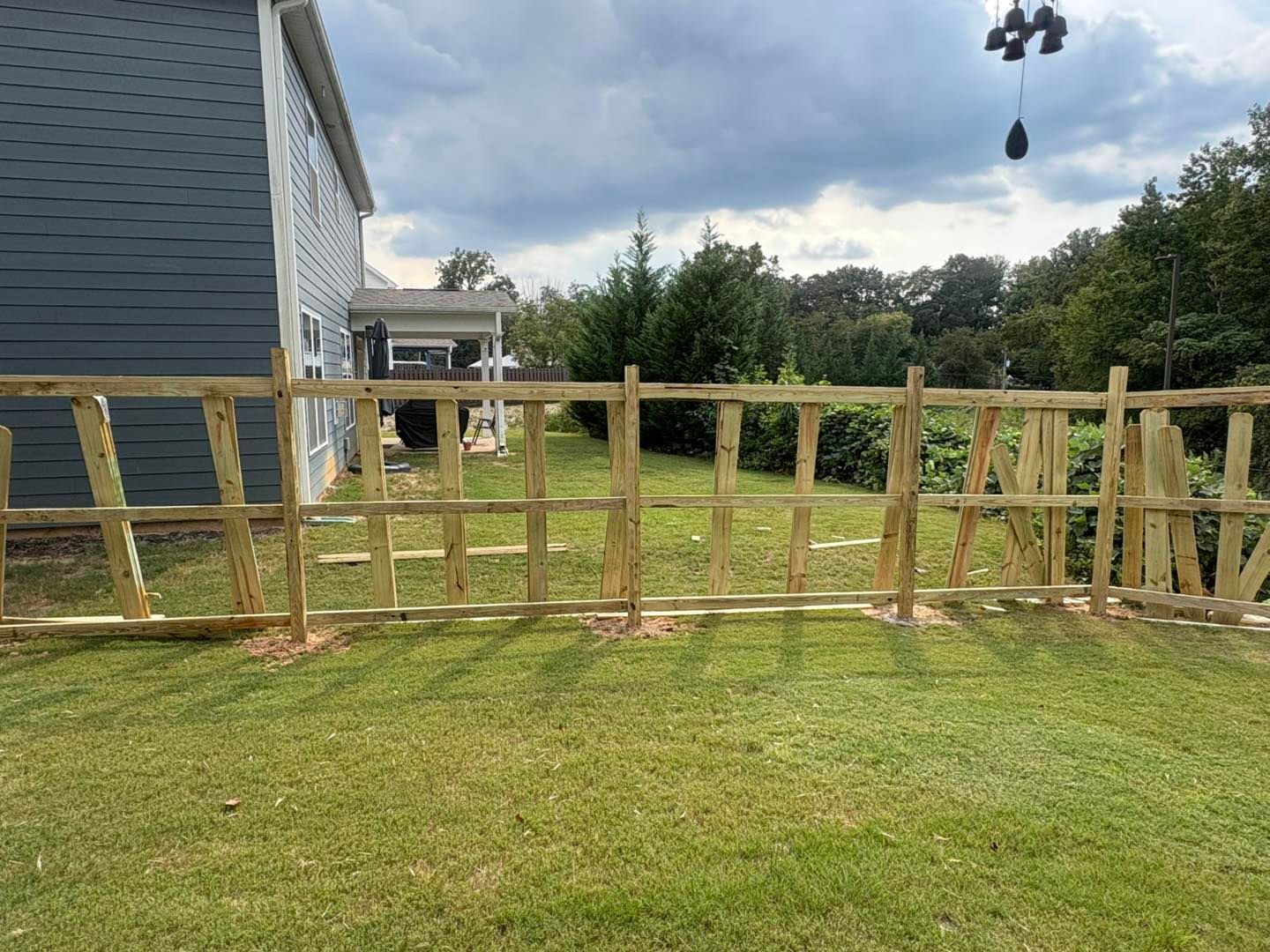 Wooden fence under construction beside a blue house, with a grassy yard and cloudy sky.