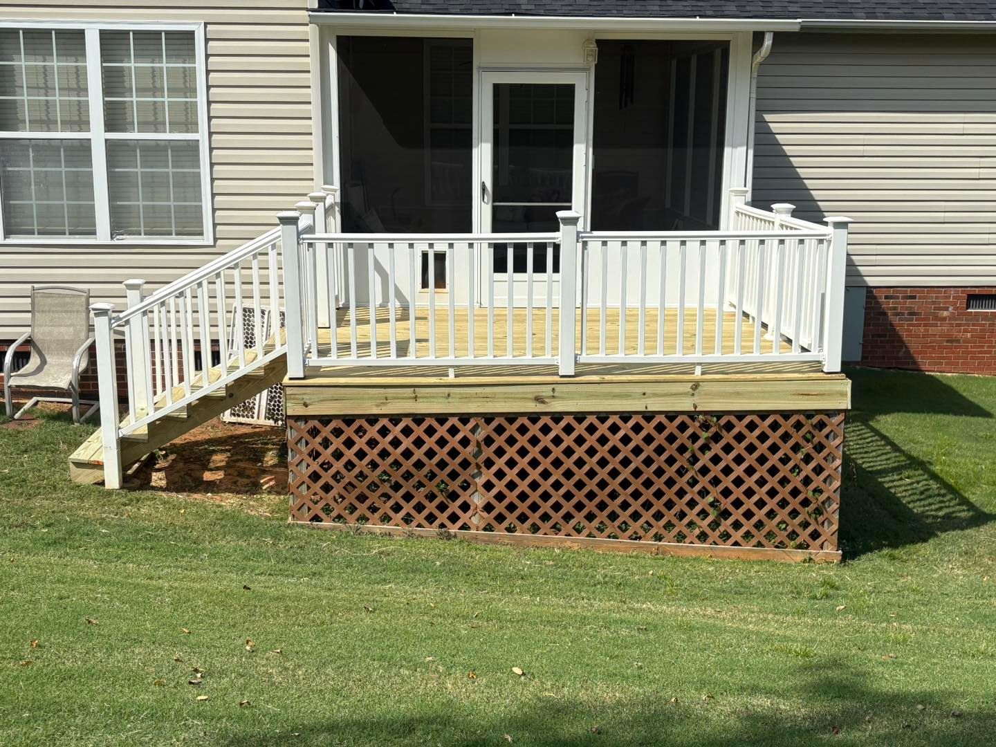 Back deck with white railing and stairs attached to a beige house, above a lattice skirt over a brick base.