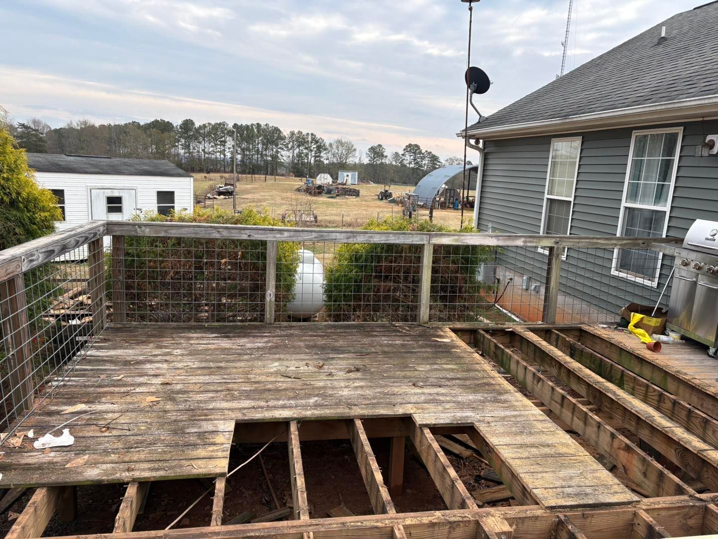 Partially demolished wooden deck beside a gray house, with exposed joists and a fenced yard beyond.