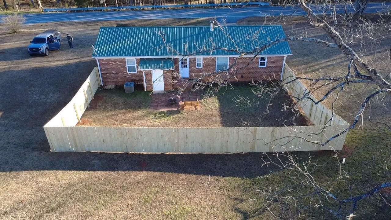 Aerial view of a brick house with a blue roof and fenced yard, surrounded by bare trees.