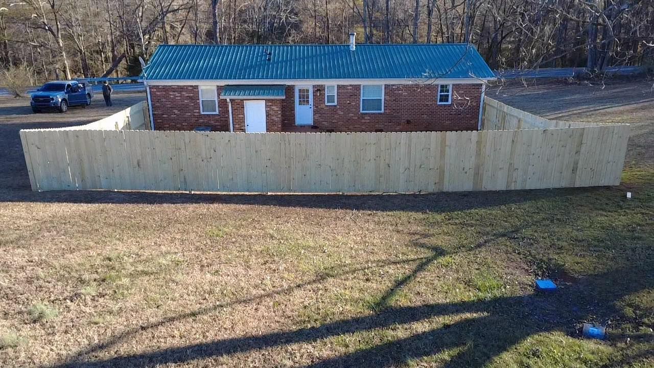 Small brick house with blue roof behind a tall wooden fence on a grassy slope.
