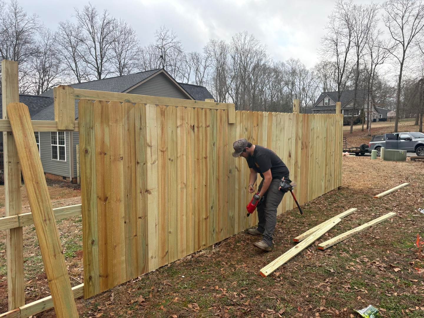 Worker nailing vertical wooden fence boards beside a house in a leaf-covered yard