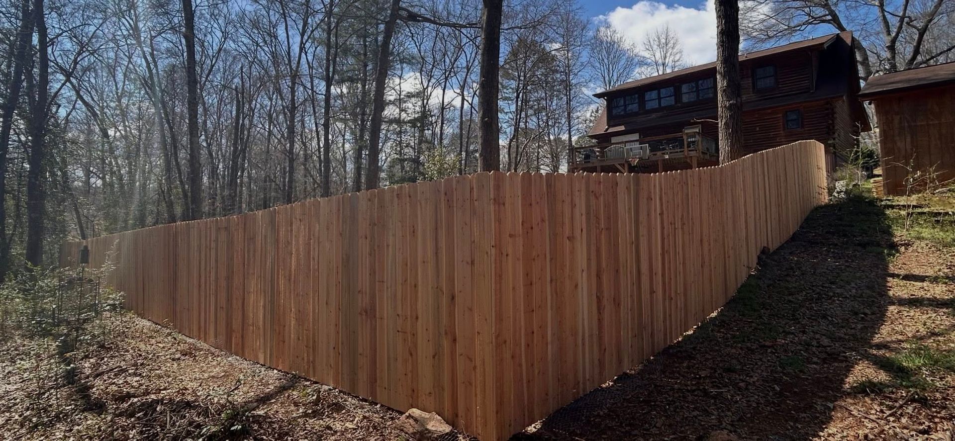 Wooden privacy fence along a wooded hillside beside a house in winter sunlight