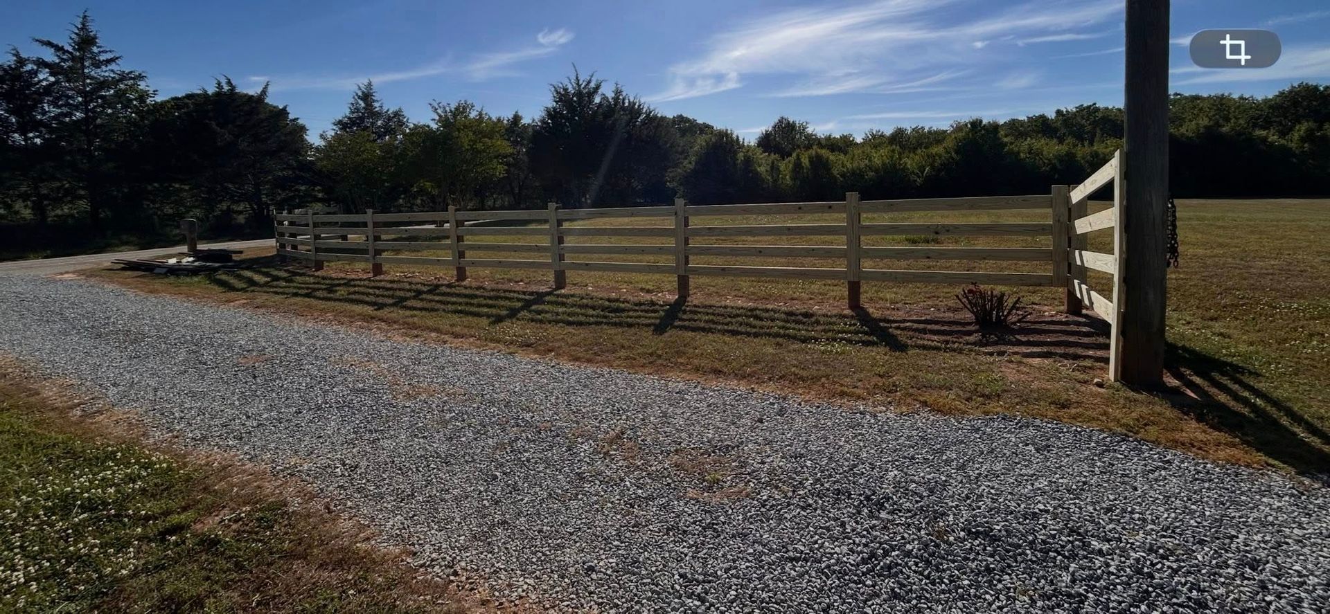 Gravel driveway beside a wooden fence on a sunny rural property with trees and open fields.