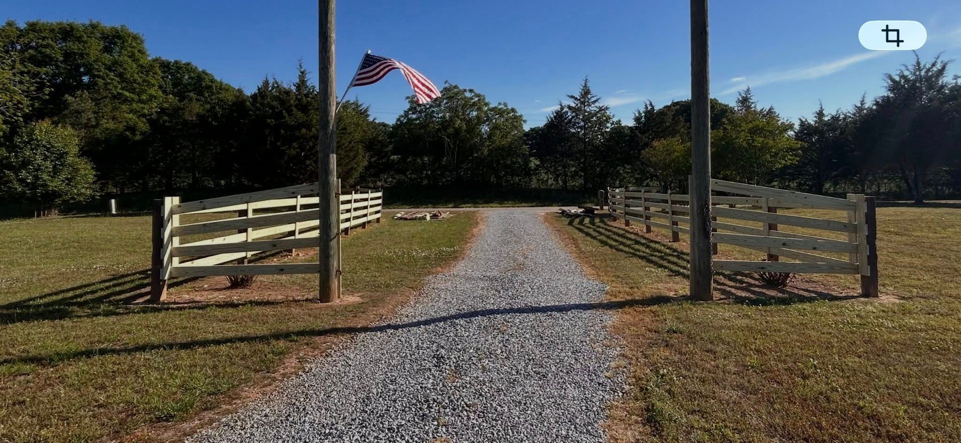 Gravel driveway through an open wooden gate with an American flag, leading to a grassy field and trees