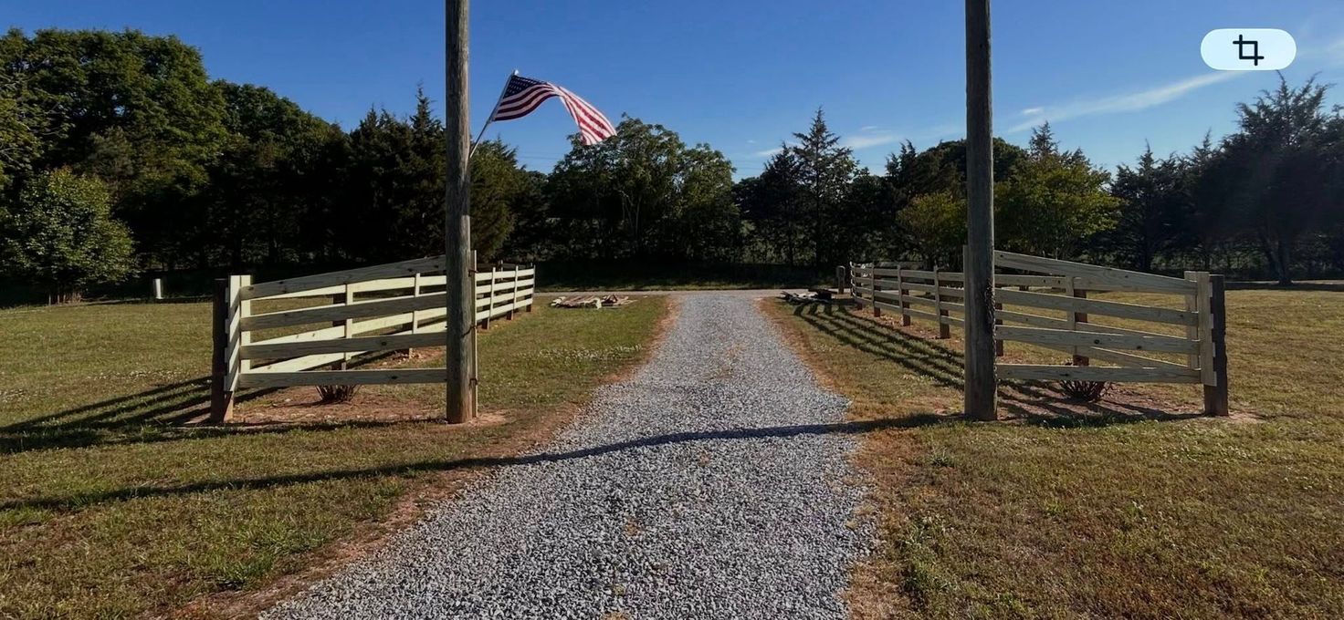 Gravel driveway through an open wooden gate with an American flag, leading to a grassy field and trees