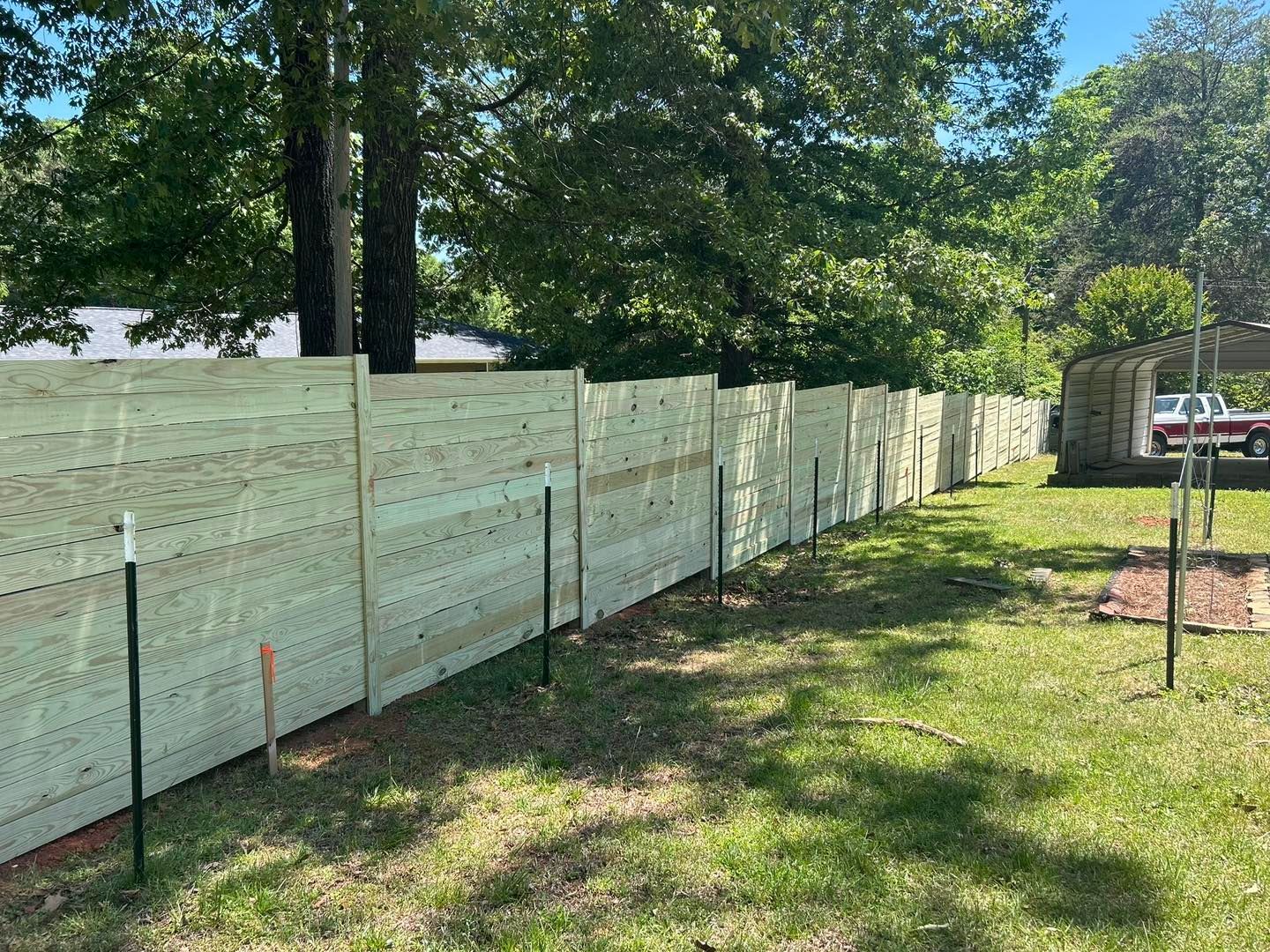 Long light-green fence panels along a grassy yard beside trees and a driveway
