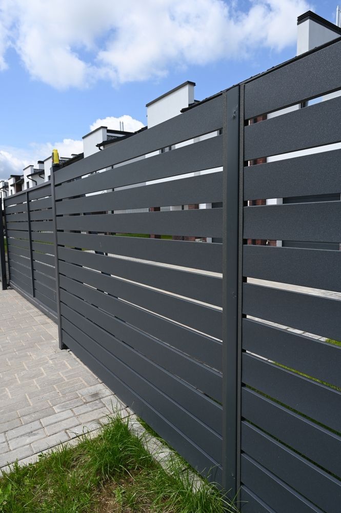 Dark gray horizontal-slat fence along a paved sidewalk under a cloudy sky