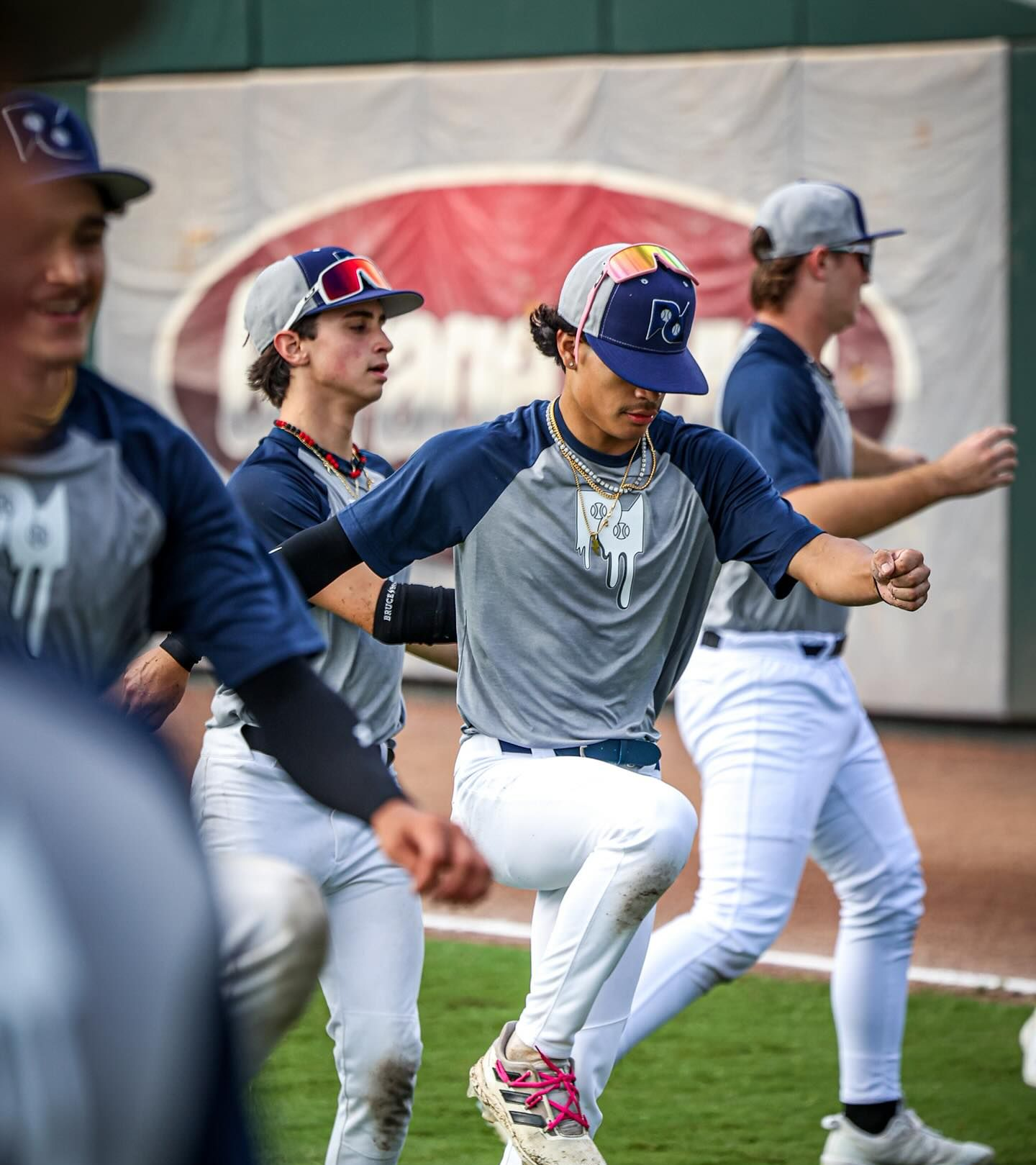 Baseball players in gray and blue uniforms stretching on a field, one with a backwards hat.