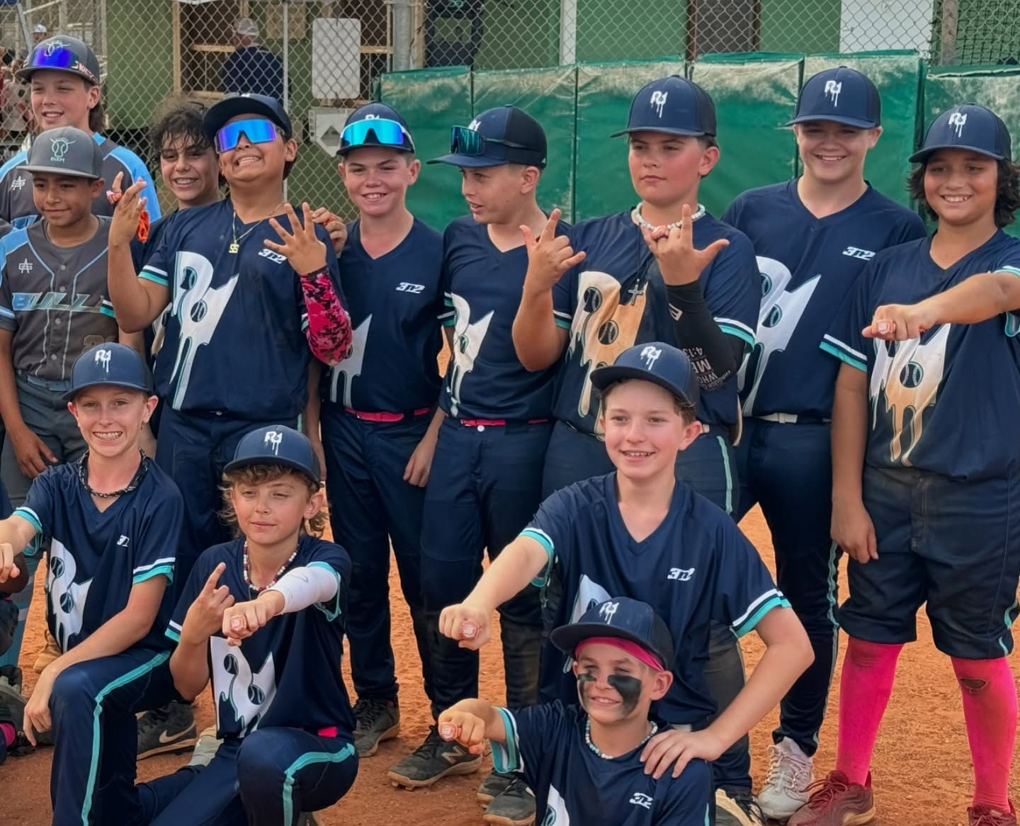Youth baseball team in dark blue uniforms pose on a field, some making hand gestures.