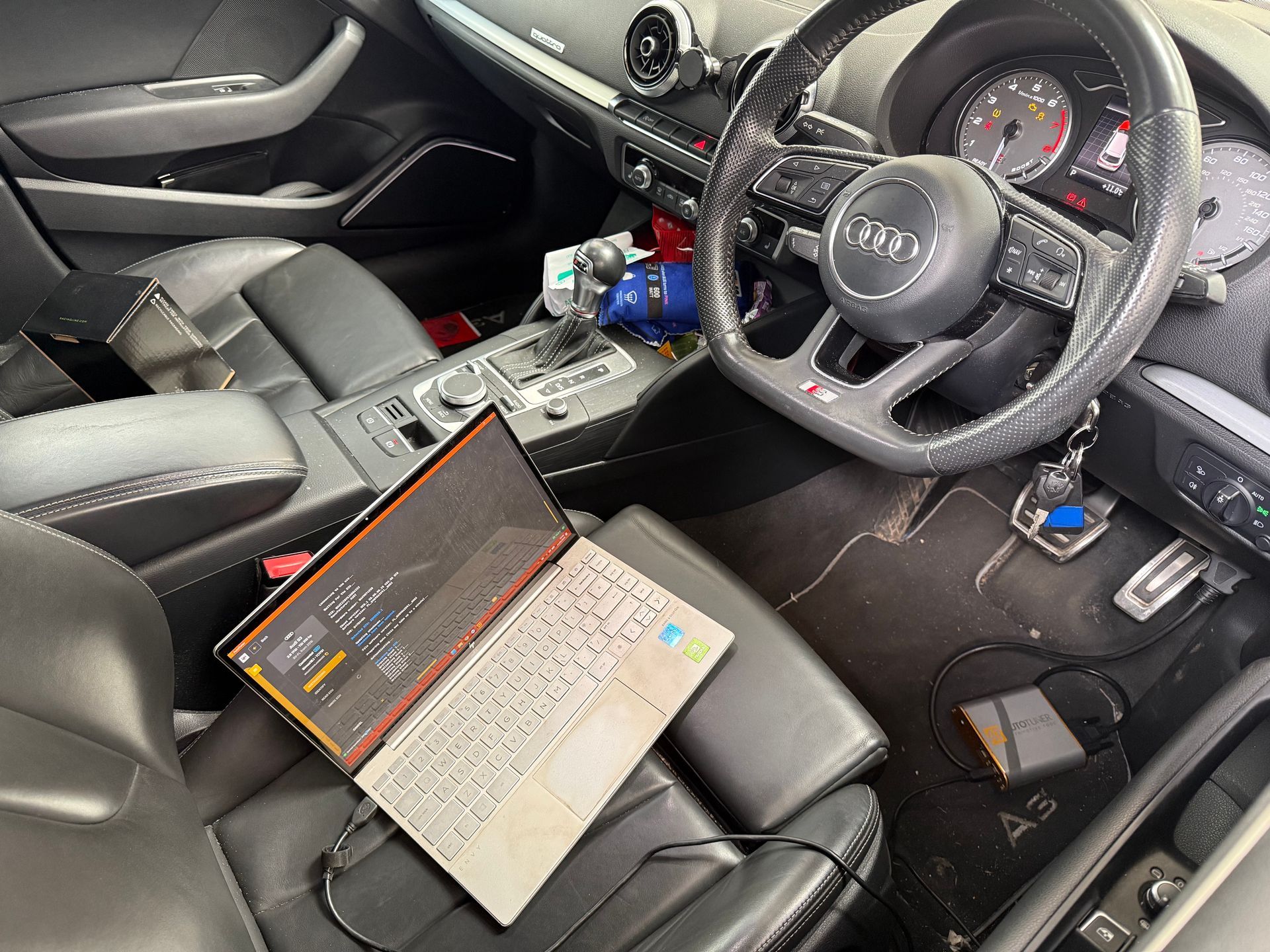 Laptop on black car seat in a right-hand-drive Audi interior with dashboard and steering wheel visible