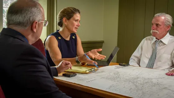 Three people in a meeting discussing papers laid out on a table, laptop, and pens are visible.