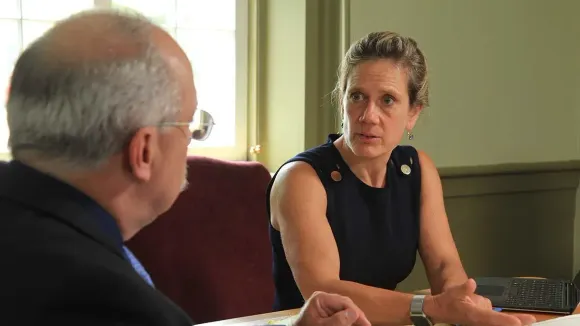 Woman in a dark sleeveless dress gestures while speaking to a man at a table indoors.