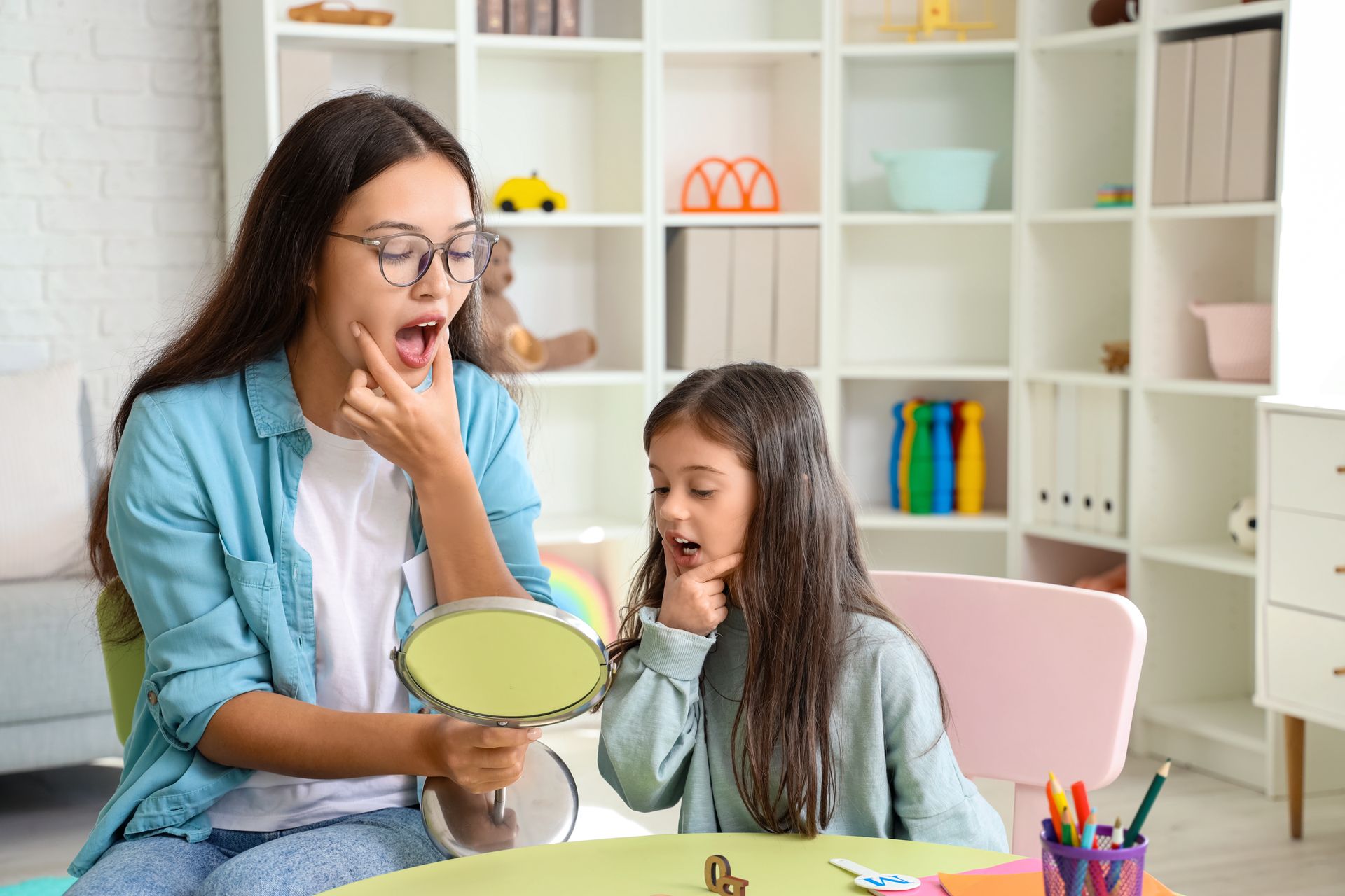 A woman is teaching a little girl how to speak in front of a mirror.