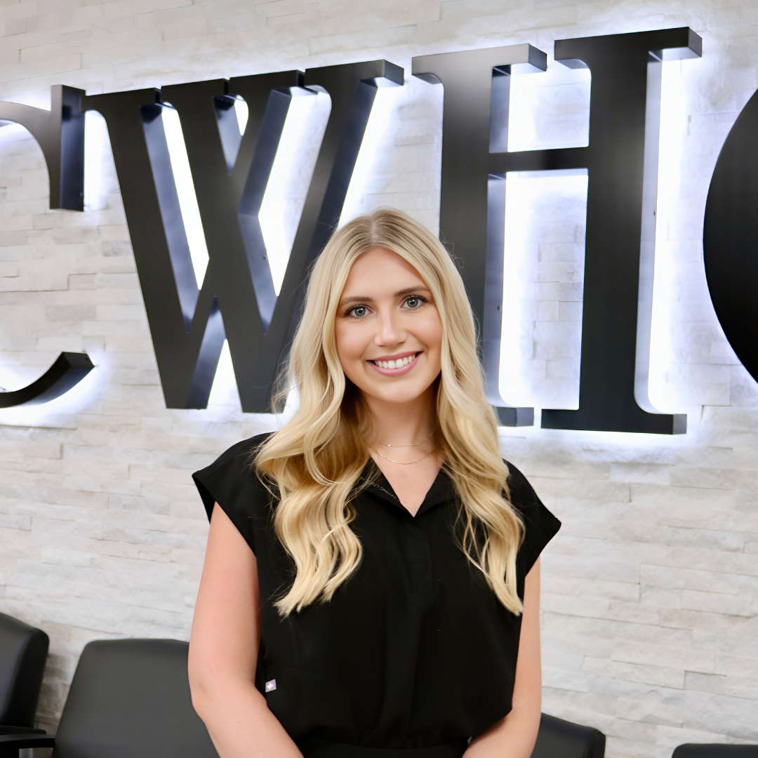 Blonde woman in a black blouse smiling in front of a wall with large backlit letters.