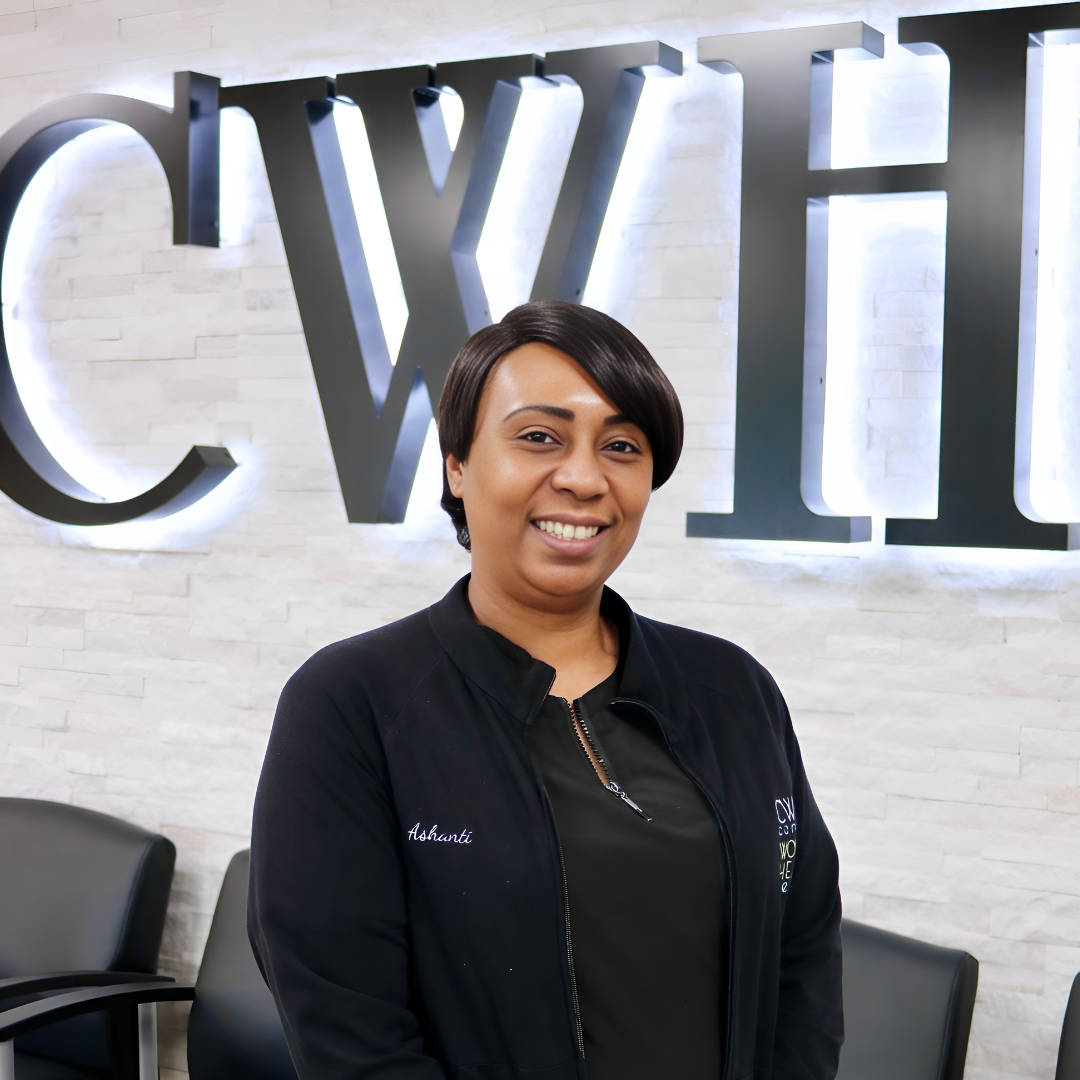 Smiling person in a black jacket standing indoors in front of large silver letters on a white wall