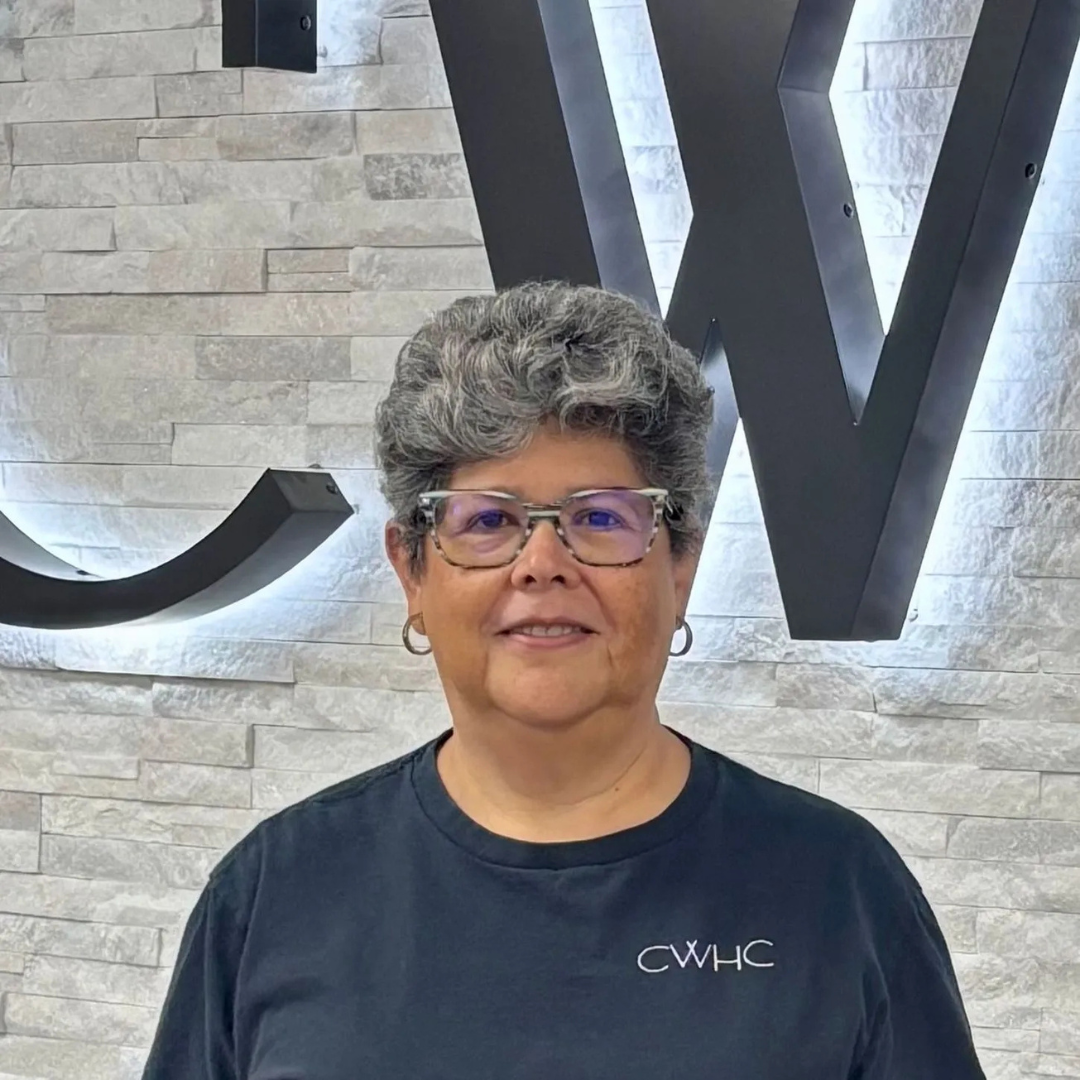 Woman with short gray hair and purple glasses standing before a lit stone wall with large letters.