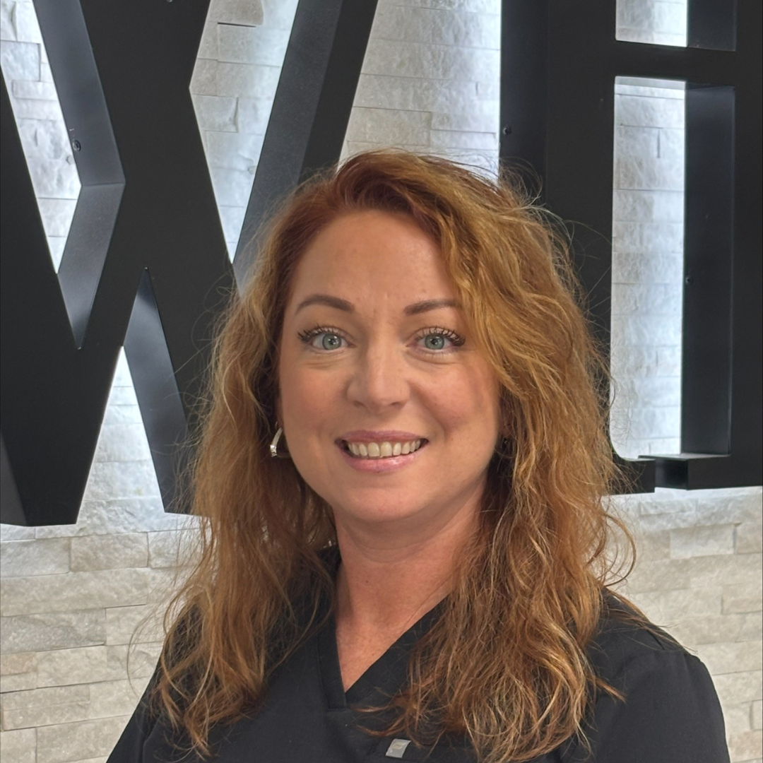 Smiling woman with curly red hair standing indoors in front of a wall sign.