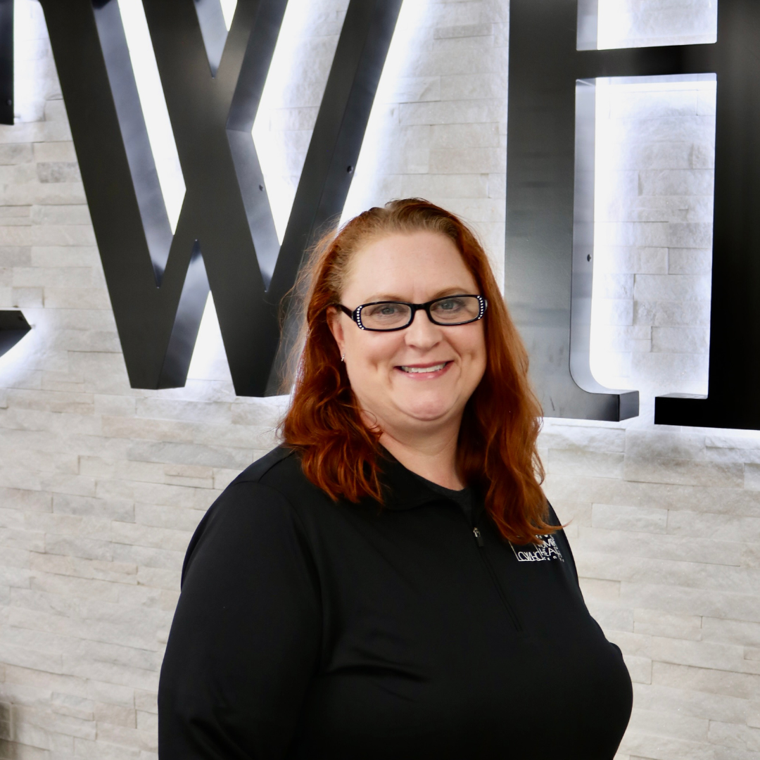 Smiling person with red hair and glasses in a black shirt standing in front of large wall letters