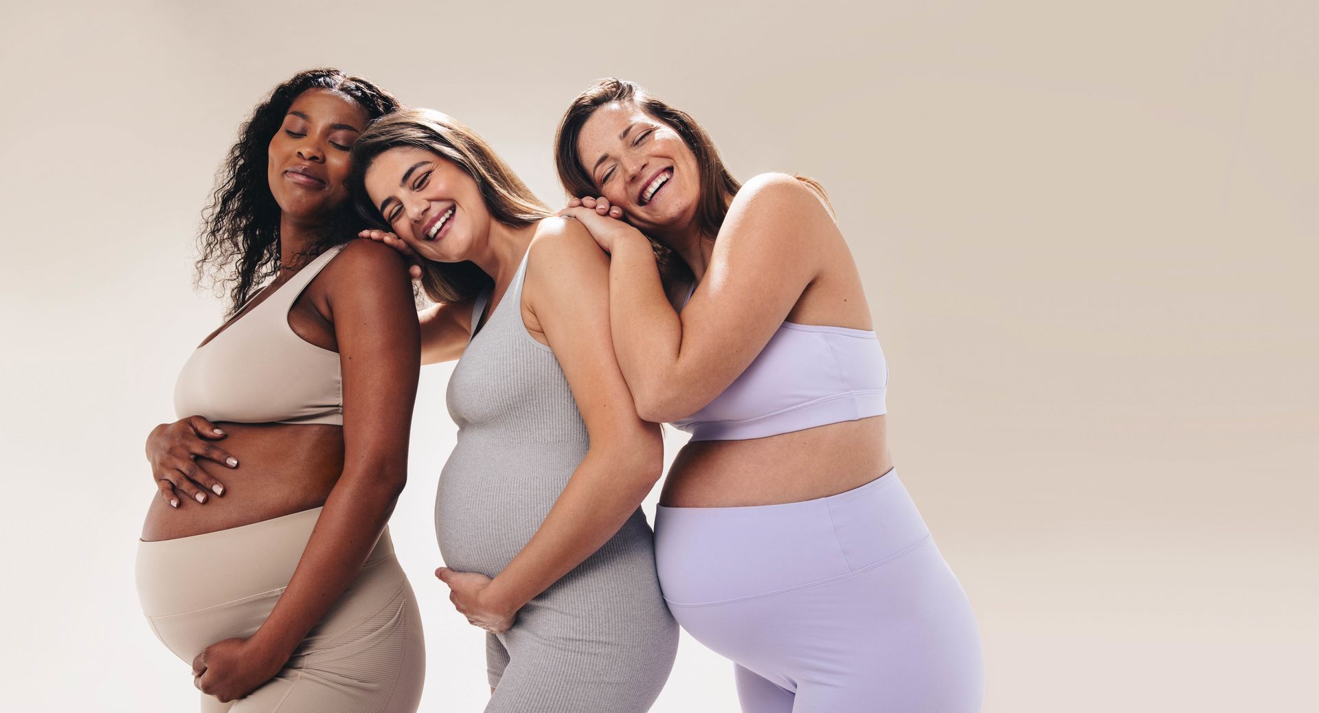 Three pregnant people in pastel outfits posing closely together against a beige background