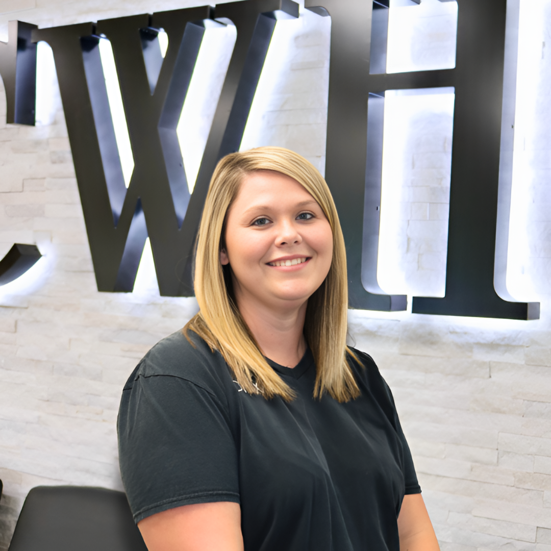 Woman smiling in a black shirt, standing before a lit office wall sign with large black letters.