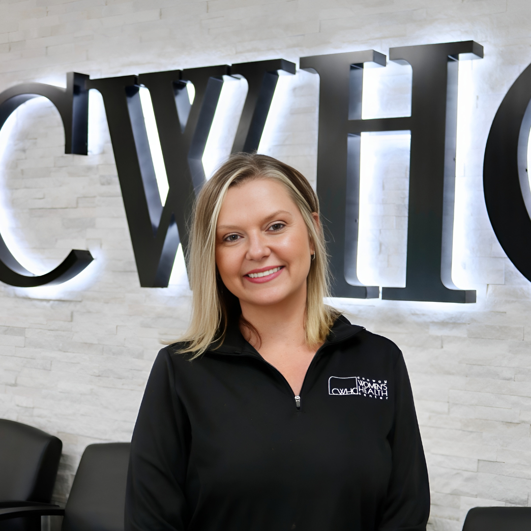 Smiling woman in black quarter-zip stands before large illuminated letters on a white brick wall.