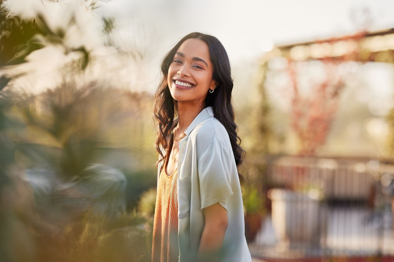Smiling woman standing outdoors in warm sunlight, with blurred greenery and buildings in the background